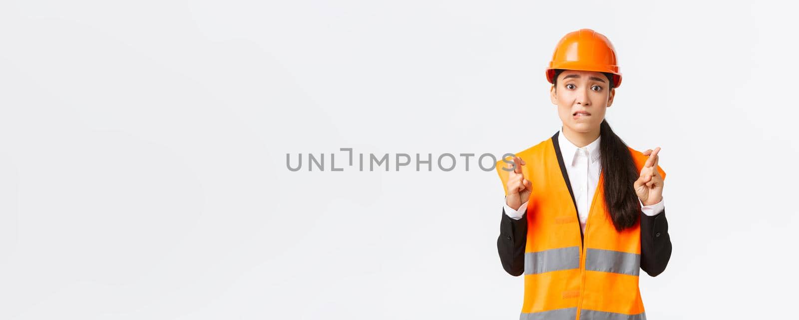 Worried hopeful asian female architect feeling nervous as waiting for inspection results at construction area, wearing safety helmet, cross fingers good luck, hope everything be well by Benzoix