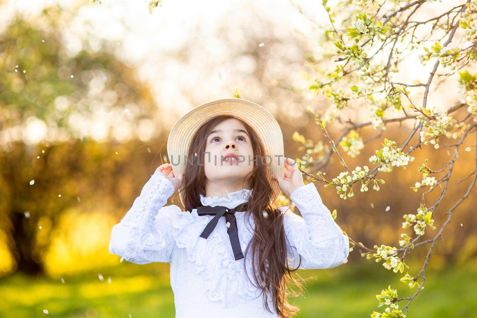 A beautiful girl in a white dress and hat, stands in the park, under flowering trees, in spring, looks up