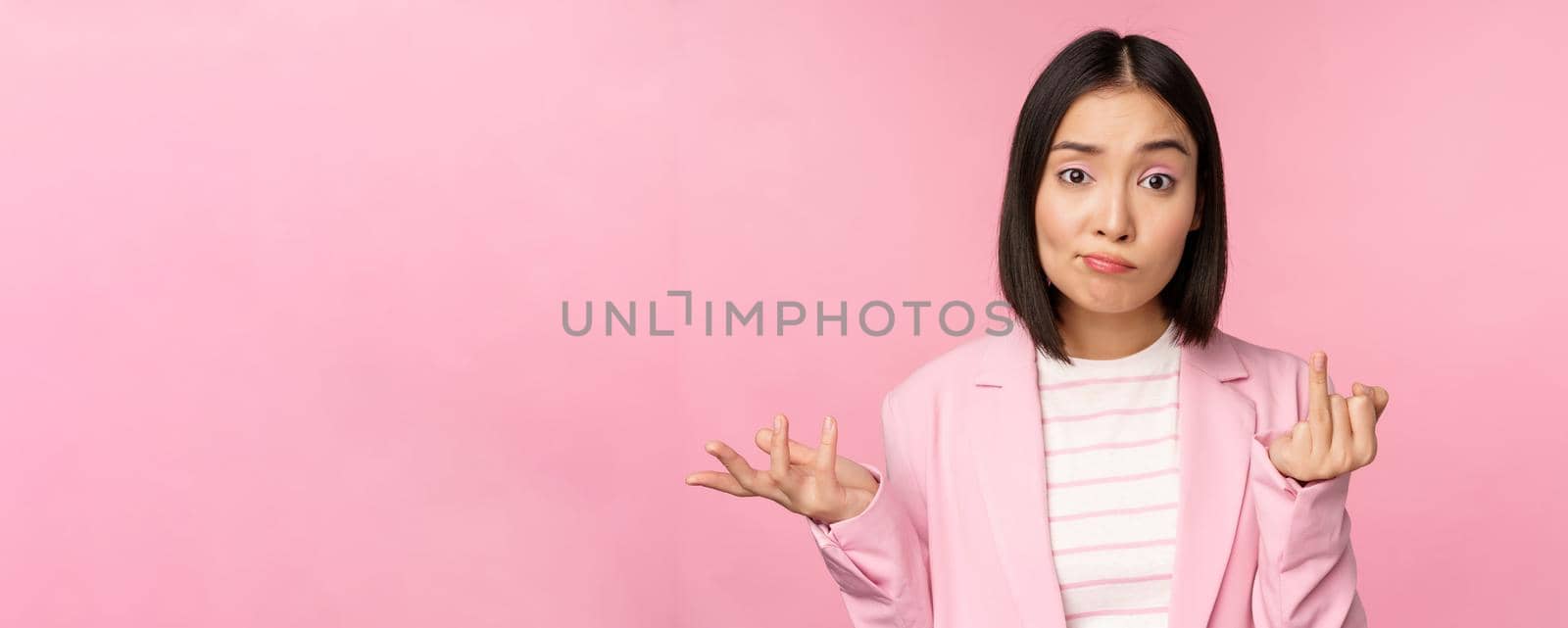 Portrait of young asian business woman, saleswoman shrugging shoulders and looking confused, clueless of smth, standing over pink background by Benzoix
