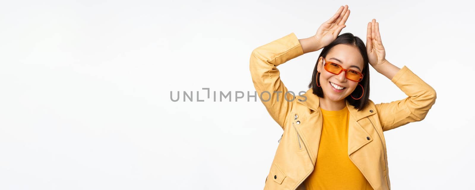 Portrait of beautiful asian girl in sunglasses, showing bunny ears hands gesture and smiling, standing happy over white background.