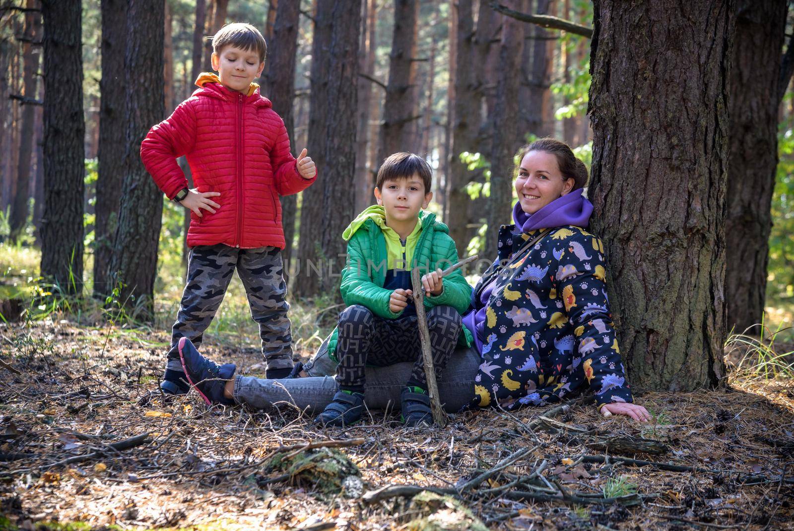 Family with mother and two children boy sibling brothers sits in the forest on felled logs or grass in summer. Happy family in colorful clothes have a hike in autumn forest.