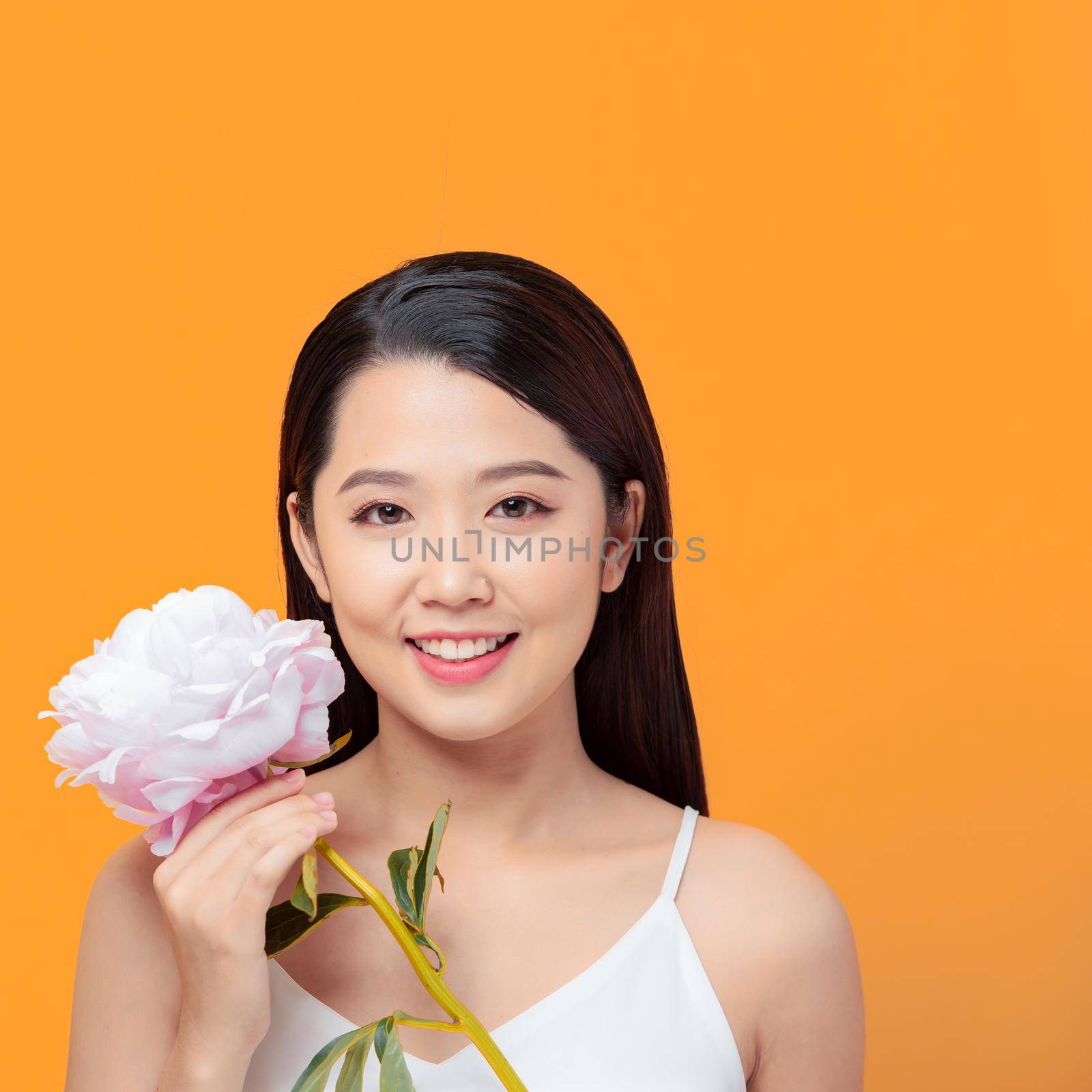 Young woman holding pink peony flower, smiling on yellow background