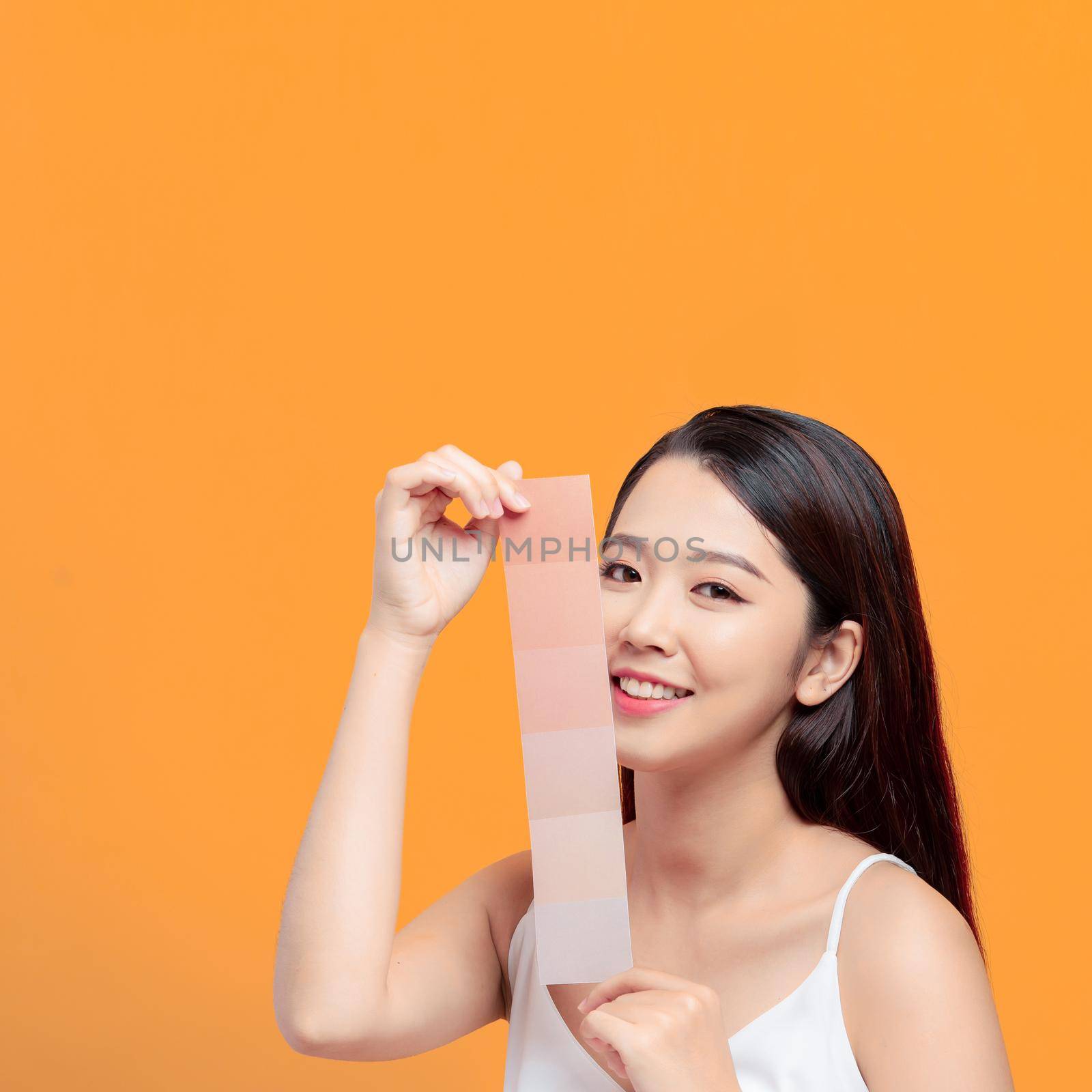 Portrait of beautiful young woman holding paper of different tones of skin on yellow background