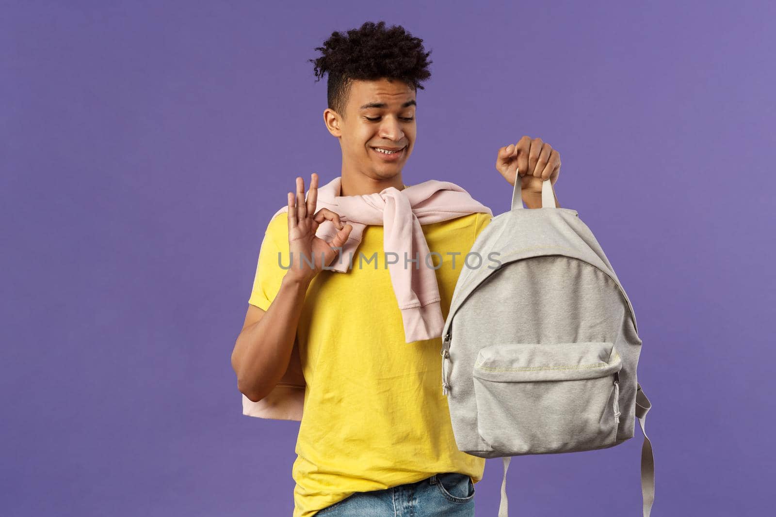 Portrait of young awkward guy, student receive gift new backpack from mom, being displeased with it grimacing but say okay, not bad, looking at rucksack standing purple background.