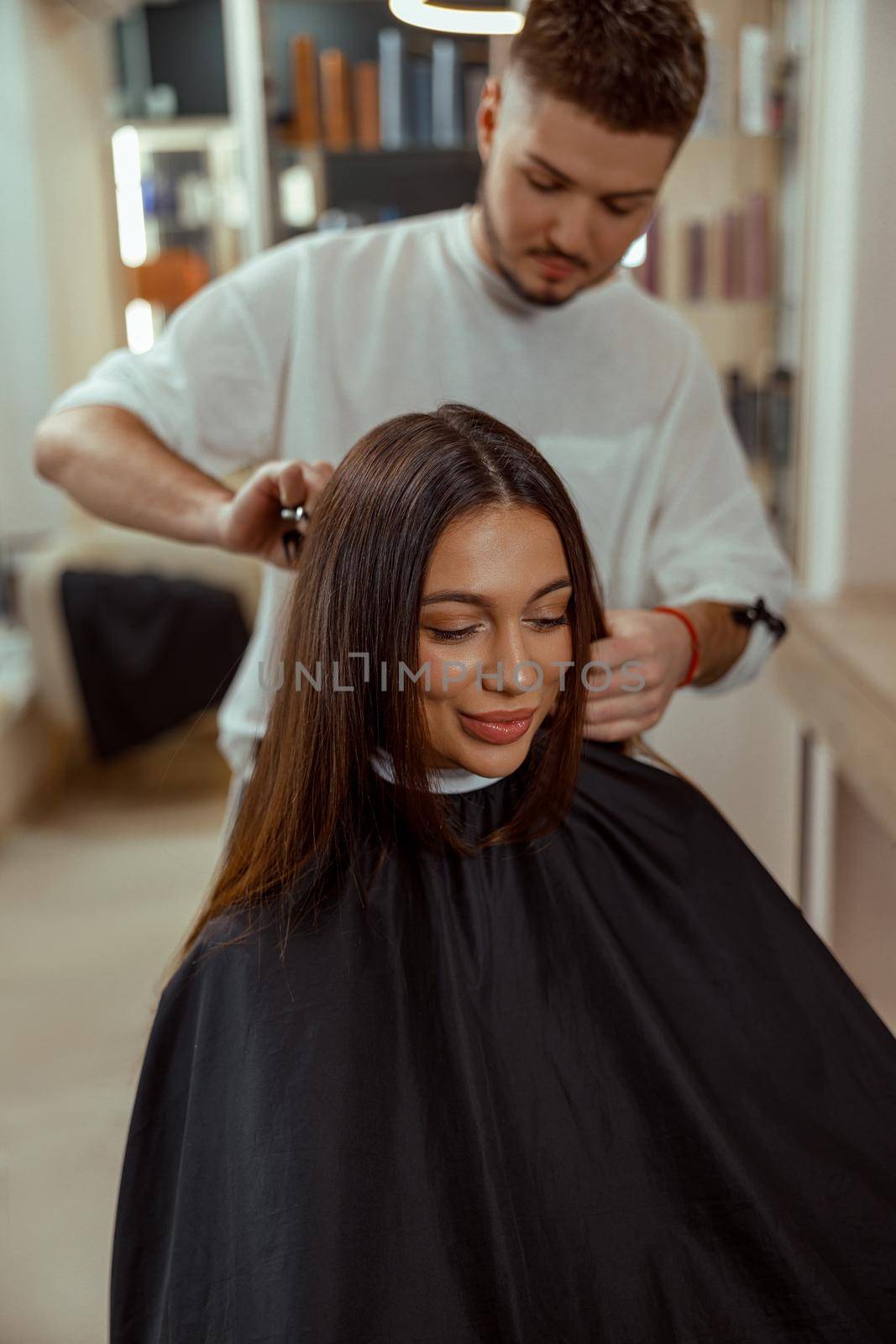 A happy young woman sits in a chair covered with a cape while a hair master cuts her hair by Yaroslav_astakhov