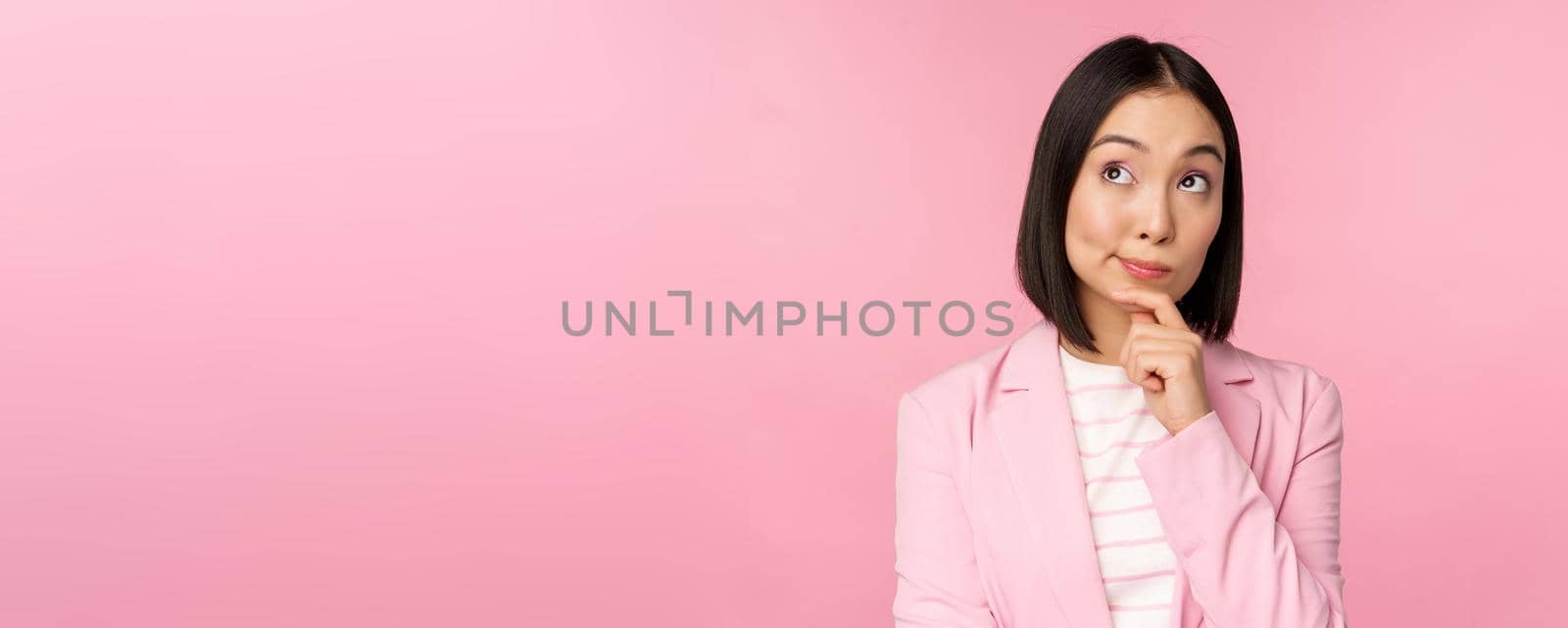 Image of asian businesswoman standing in thinking pose, brainstoming, wearing suit. Korean saleswoman, entrepreneur posing against pink background by Benzoix