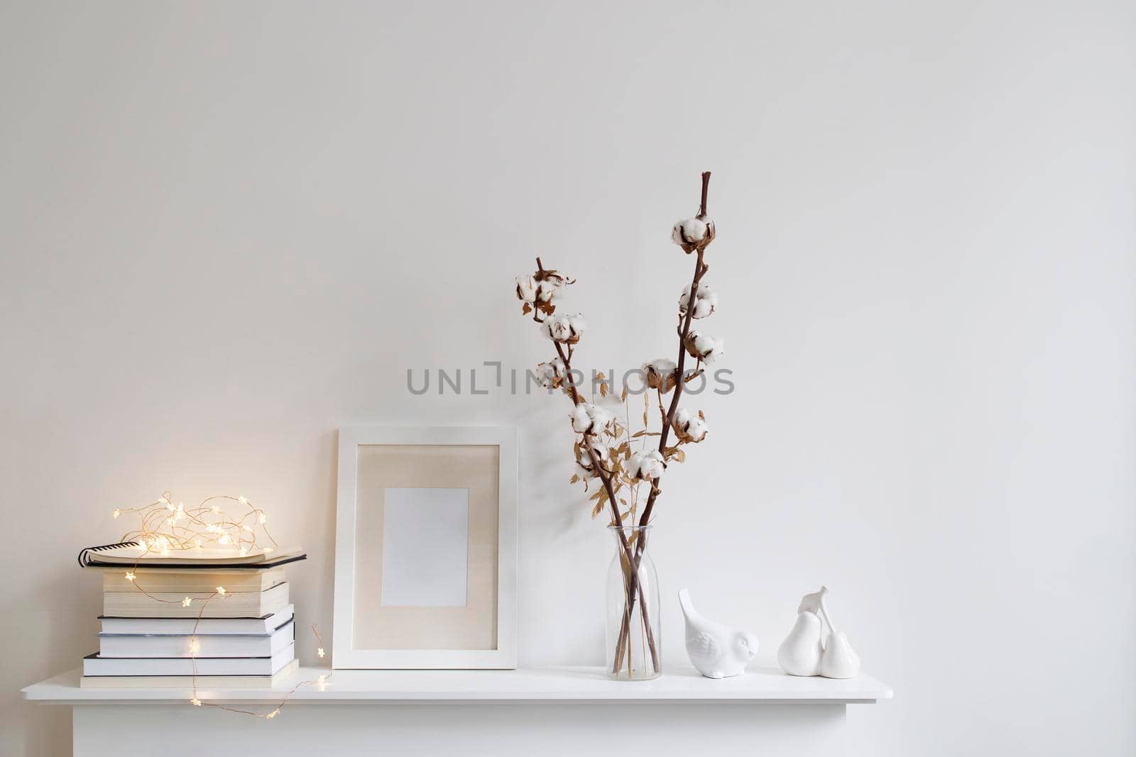 Scandinavian style room interior in white tones. A vase with cotton flowers, a stack of books, a photo frame, a bird figurine on a wooden surface of a shelf. Copy space. by elenarostunova