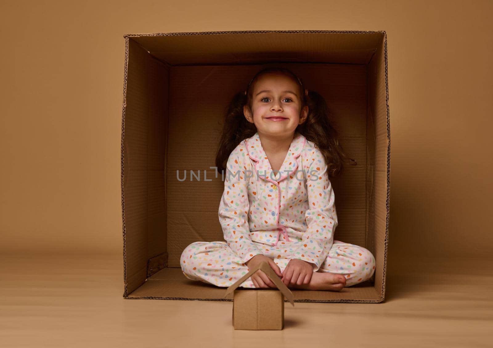 Adorable little girl sitting inside a cardboard box with a craft house model, smiles looking at camera, posing against beige background with copy ad space. Concept of housing, insurance, real estate by artgf