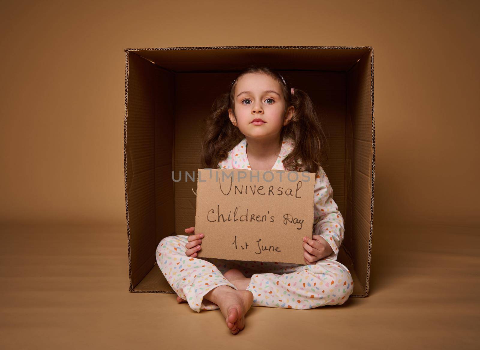 International Children's Day. Social advertising, information campaign for the fight for children's rights. Cute little girl with two ponytails holding cardboard poster sitting inside a cardboard box.