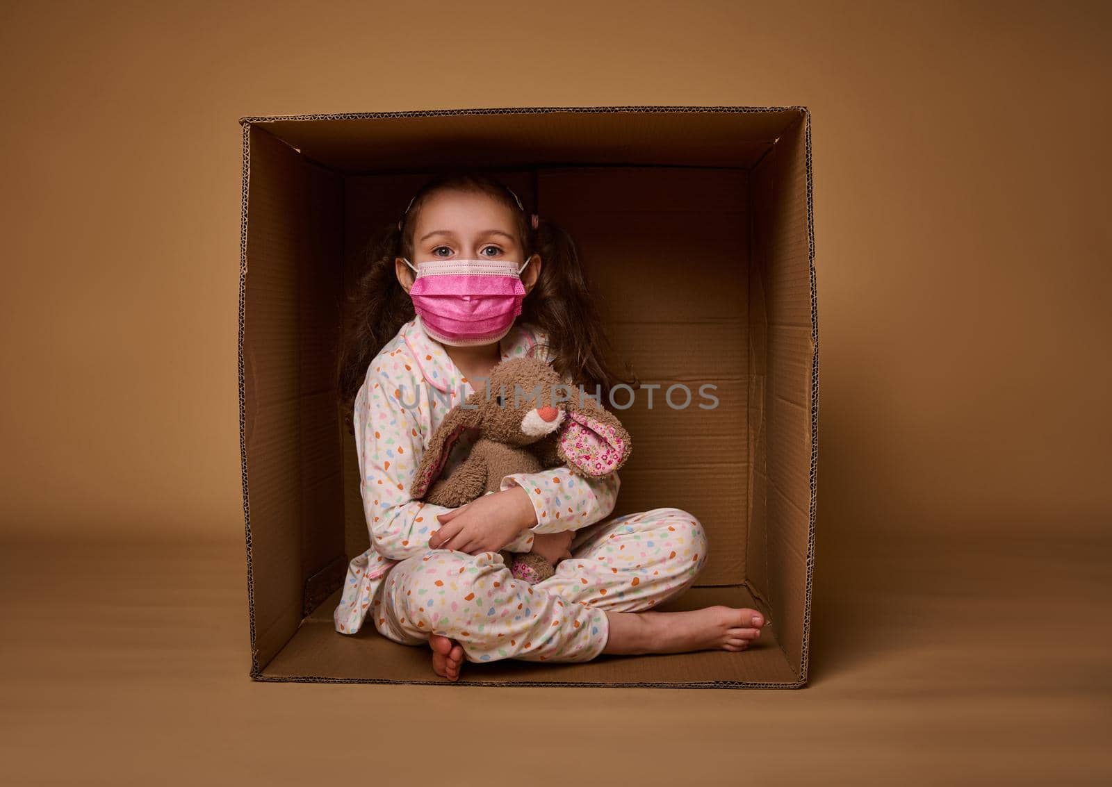 European baby girl in pajamas wearing a pink medical protective mask hugs her plush rabbit toy, sitting inside a cardboard box, looking at camera, isolated over beige background with copy ad space