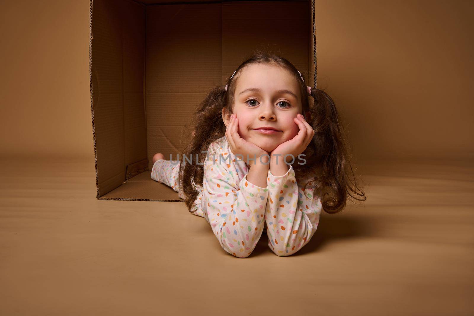 Smiling beautiful positive pleasant baby girlin pajamas with two ponytails inside a cardboard box, looking at camera isolated over beige background with copy space for ads