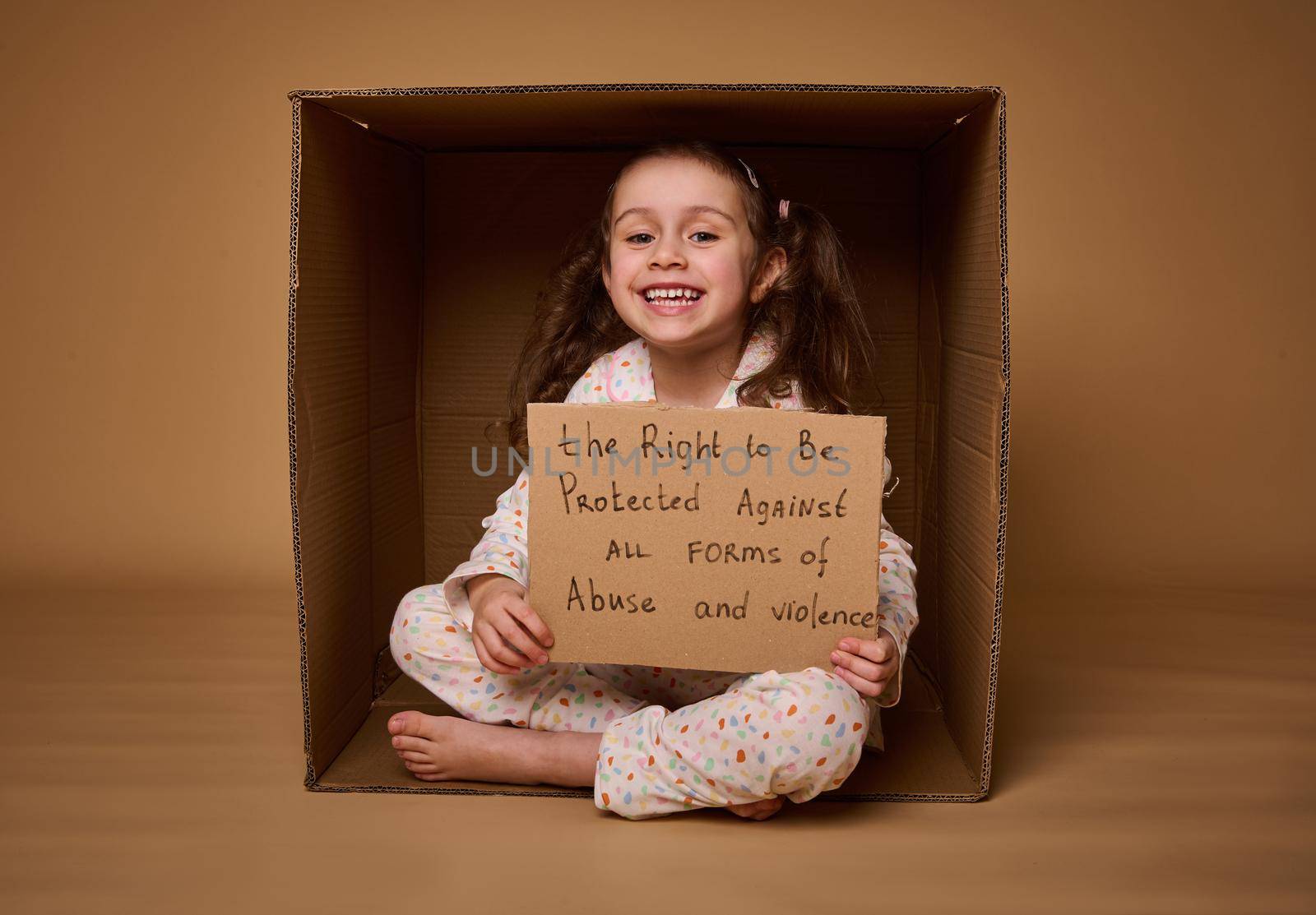 Adorable little European girl holding a poster with a message to be protected from abuse and violence, sitting inside a cardboard box. Social advertisement for International Children day. 1st July