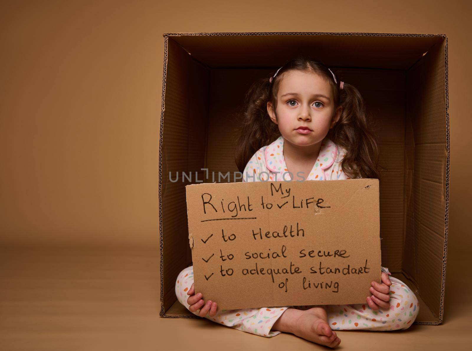 Adorable little Caucasian girl holding a cardboard poster with social message, calling for respect her rights, isolated over beige background with copy space. World Children's Day on July 1st.