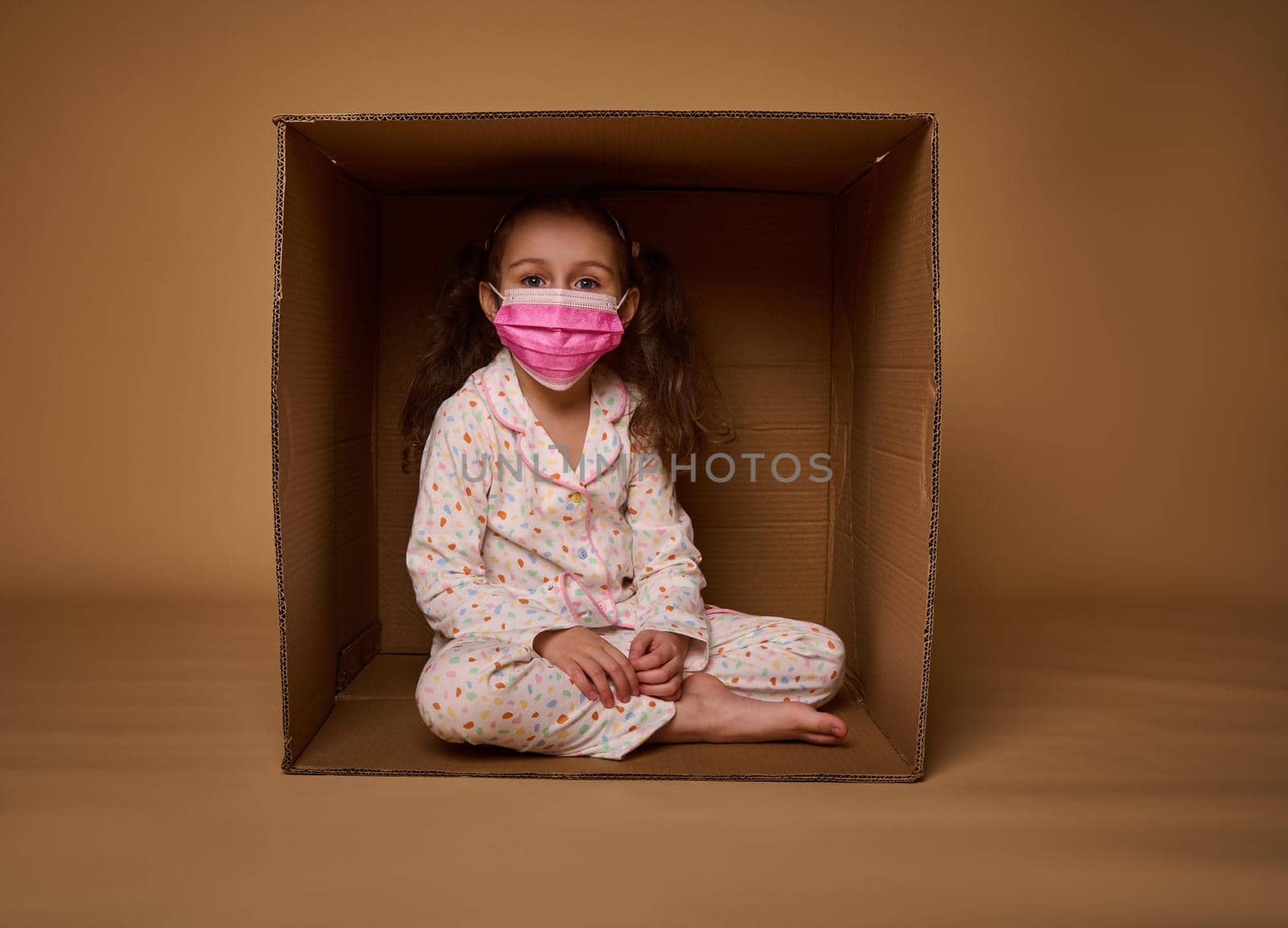 Cute little girl in pajamas wearing a pink medical protective mask sits inside a cardboard box, looks at camera, isolated over beige background with copy ad space by artgf