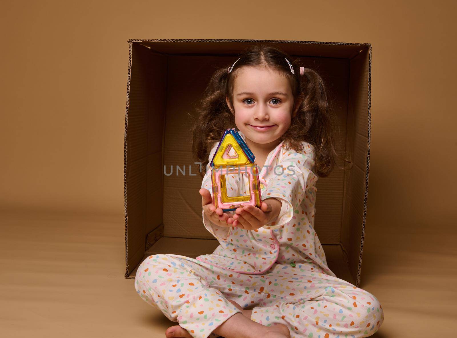 Little Caucasian girl in pajamas holding a colorful house built from magnetic construction blocks, sitting inside a cardboard box and cutely smiling looking at camera, isolated over beige background