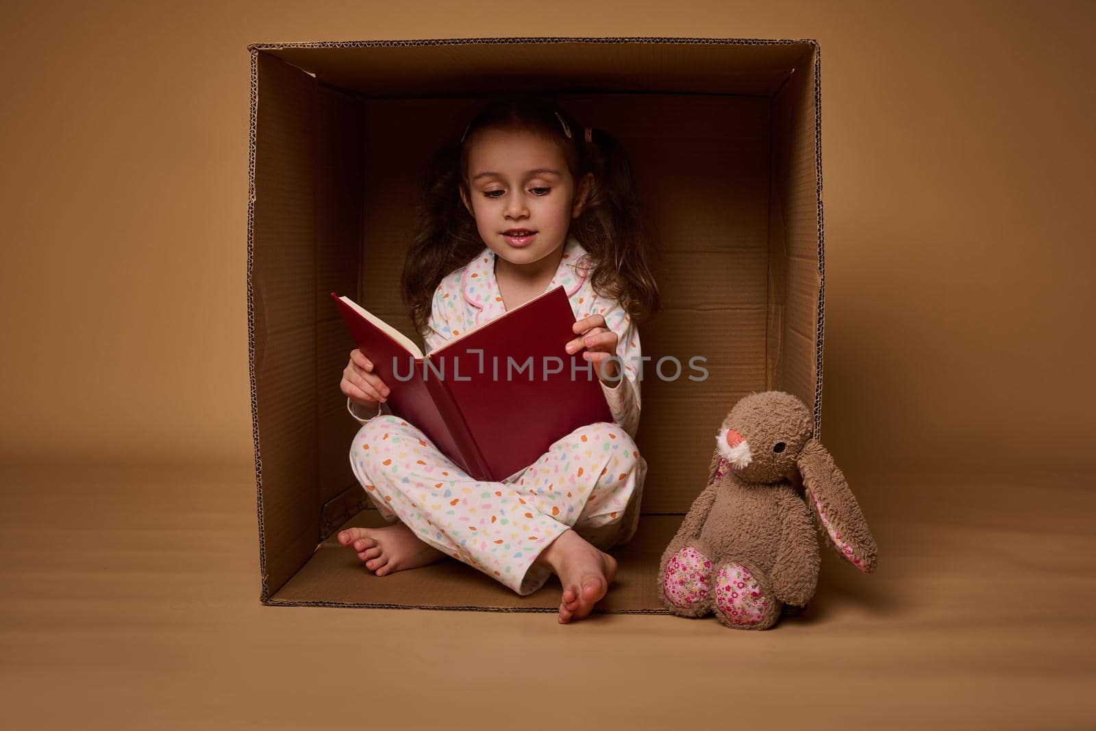 Smiling beautiful positive pleasant baby girl in pajamas with two ponytails inside a cardboard box, looking at camera isolated over beige background with copy space for ads by artgf