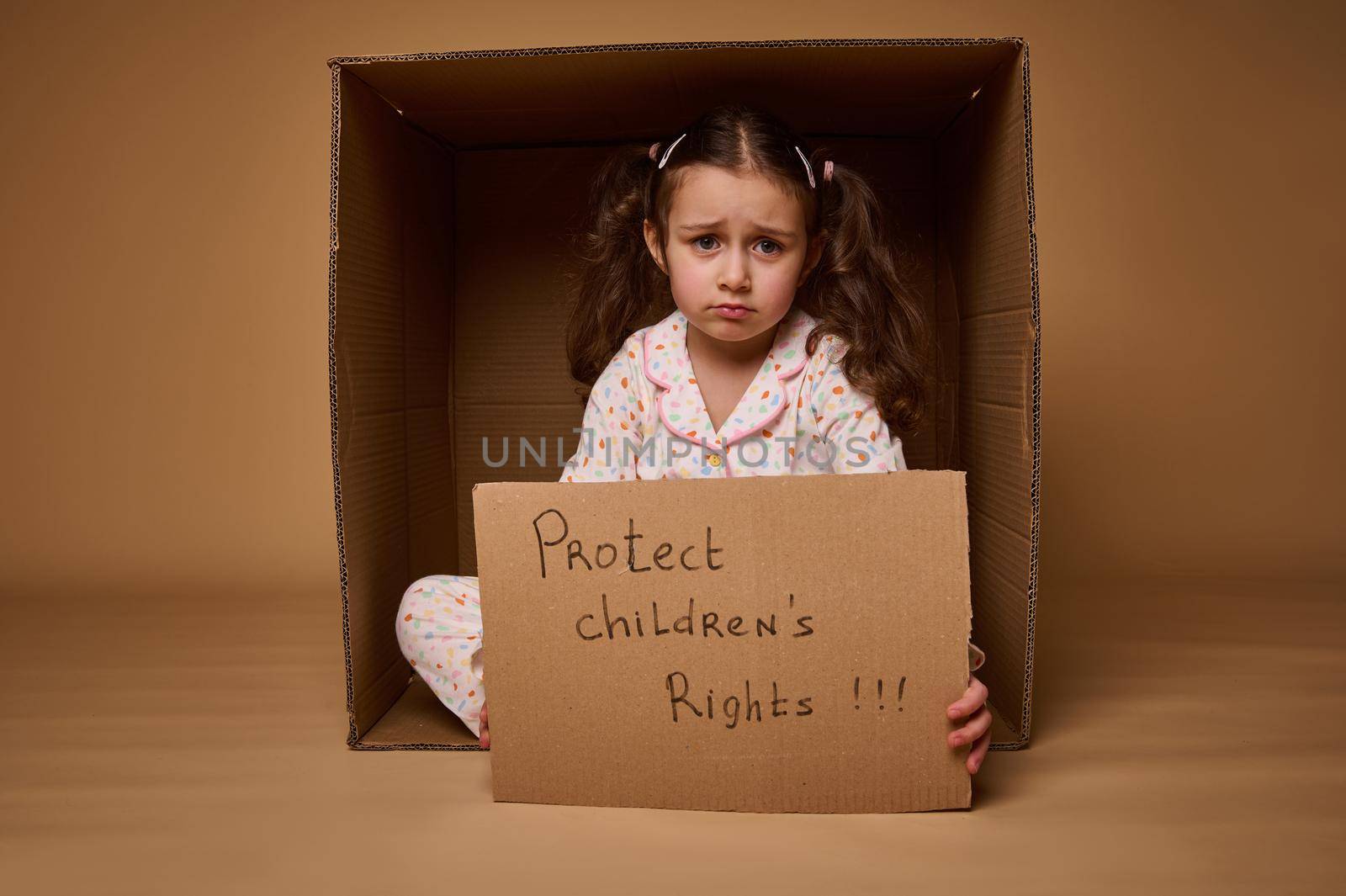 Upset little Caucasian girl holding a cardboard poster with social message, calling for respect for children's rights, isolated over beige background with copy space. World Children's Day on July 1st.