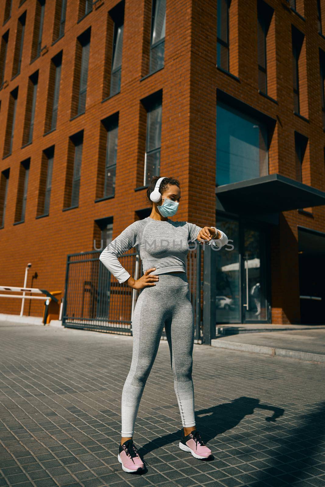 Sporty woman in headphones, mask checking time, jogging in the city. Sports concept