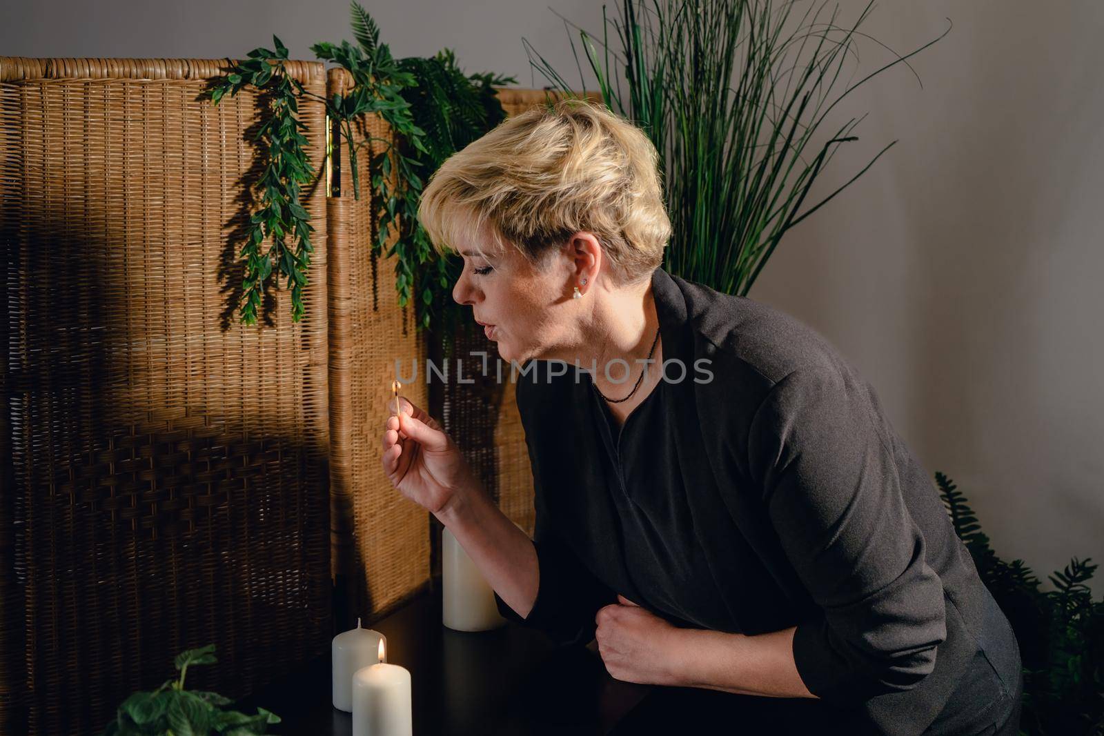 Portrait of a blonde, mature, concentrated and professional beautician, lighting scented candles with a hot wax massage matc.h. Relaxing atmosphere and soft lighting, decoration of plants and candles, massage table in the background and towels. Vertical