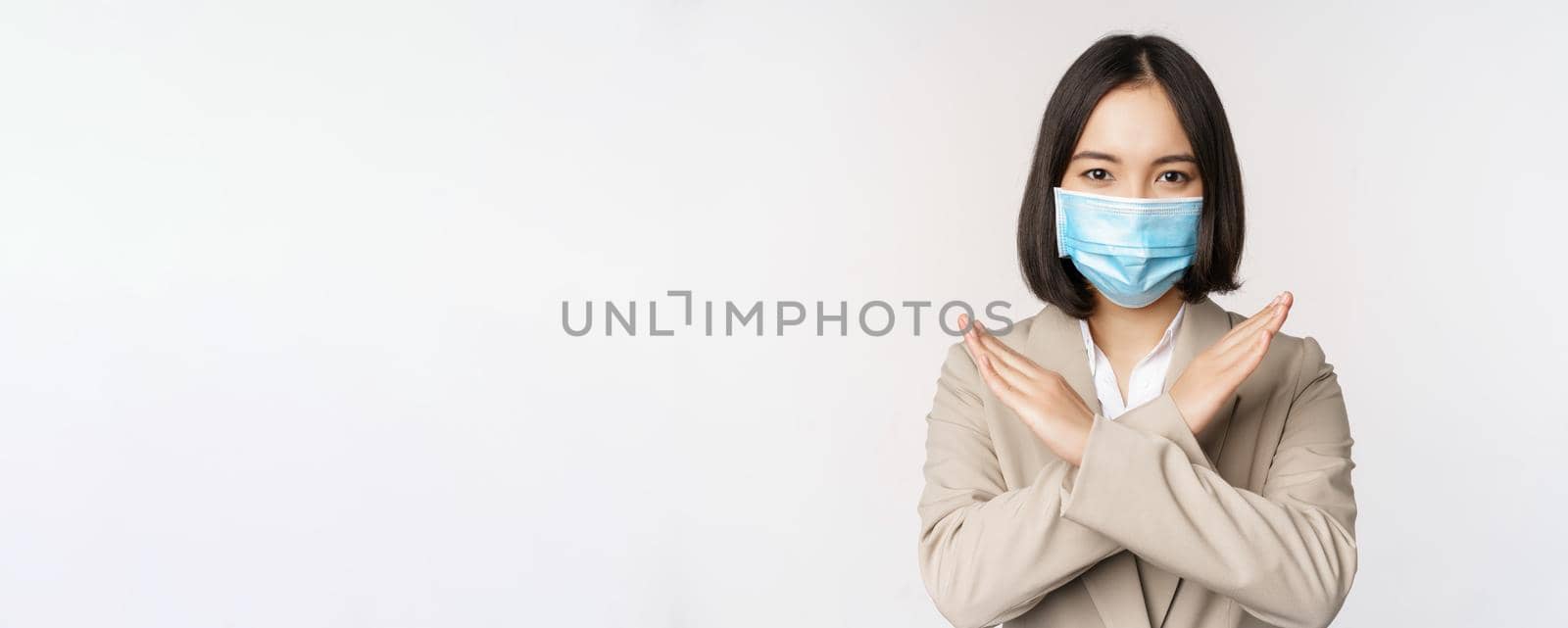 Coronavirus and workplace concept. Image of asian businesswoman, female in medical face mask shows stop, cross prohibit gesture, stands over white background.