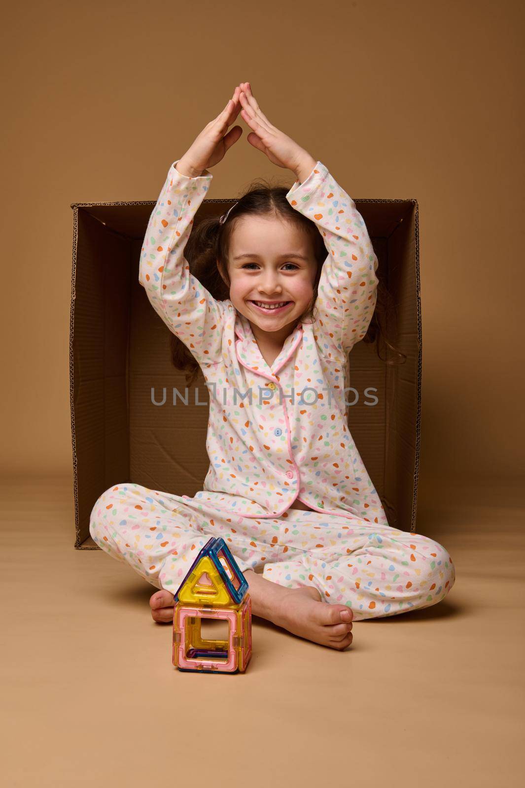 Beautiful child girl holds her hands above her head in the shape of a house roof, sitting inside a cardboard box while building house from magnetic constructor, smiles at camera over beige background