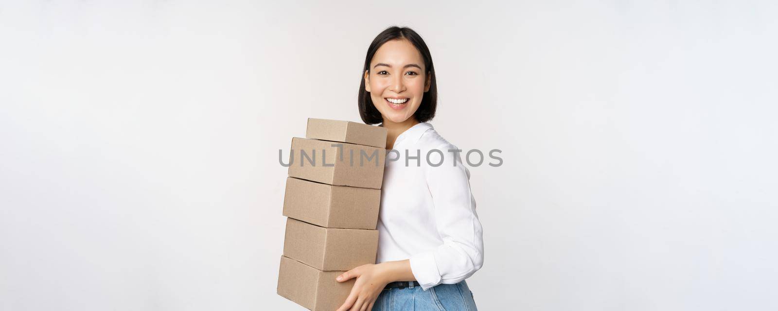 Image of smiling, happy asian woman holding pile of boxes, concept of delivery, shopping or business, white background.