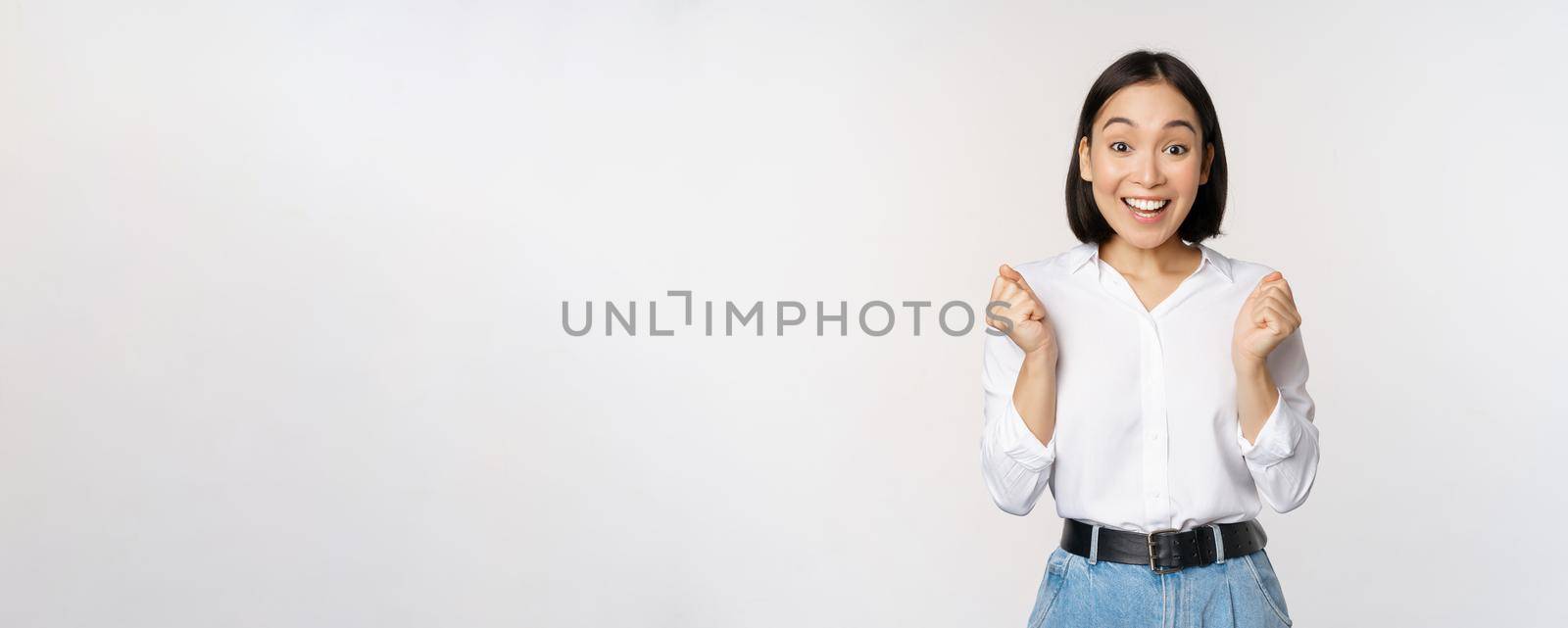 Enthusiastic asian woman rejoicing, say yes, looking happy and celebrating victory, champion dance, fist pump gesture, standing over white background.