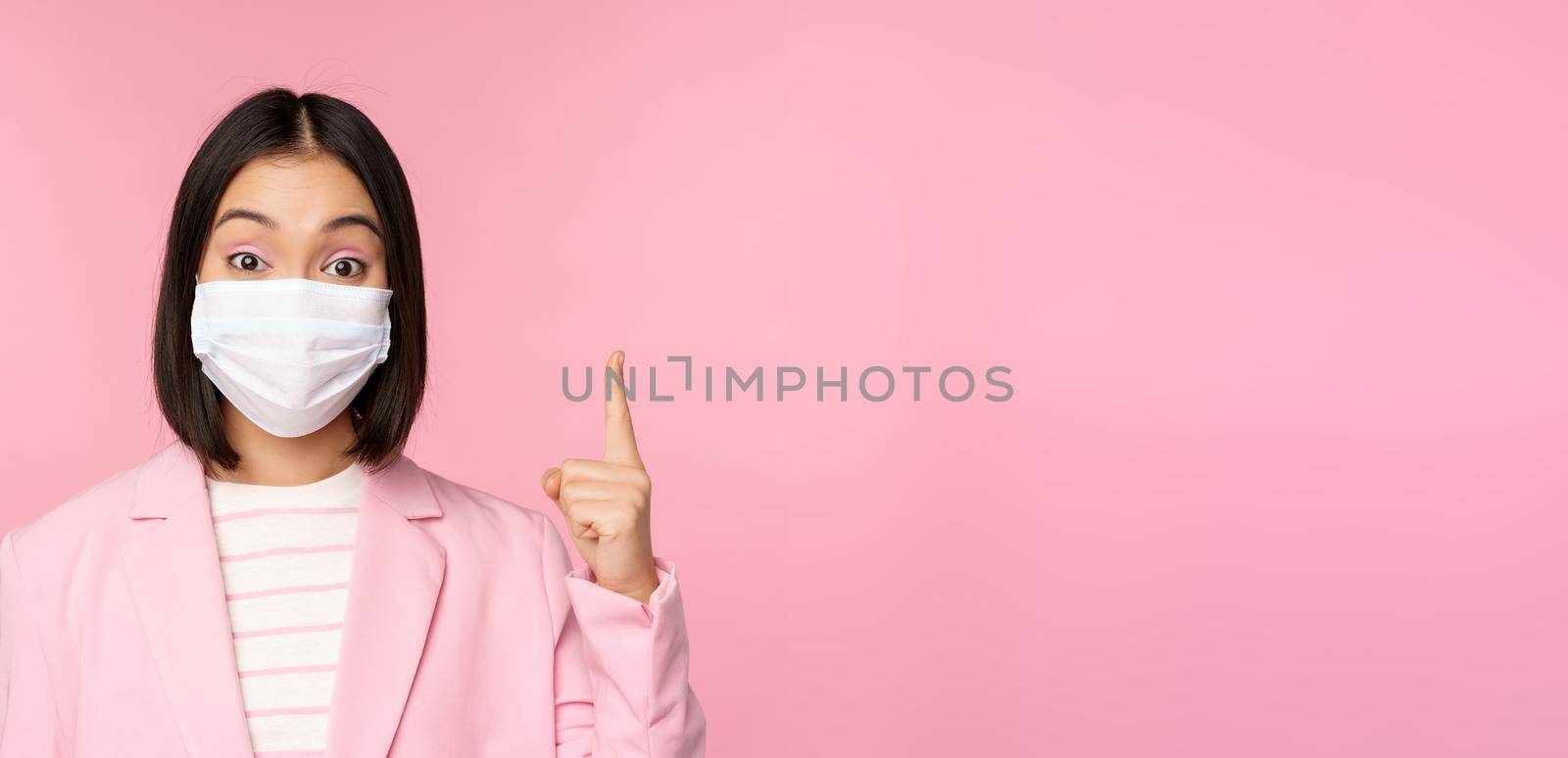 Close up portrait of asian businesswoman in medical face mask and suit, pointing finger up, showing advertisement, top banner, standing over pink background.