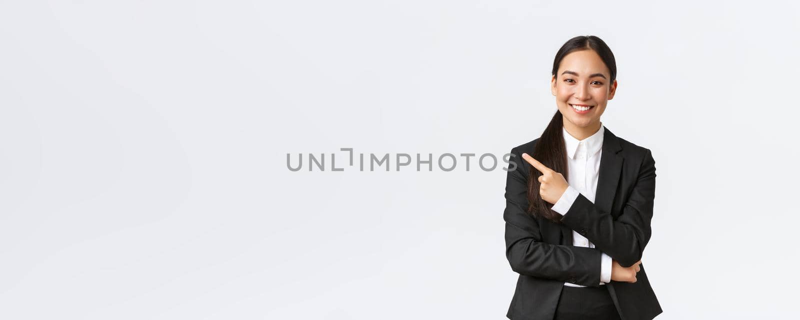 Happy professional asian female manager, businesswoman in suit showing announcement, smiling and pointing finger left at product or project banner, standing white background.