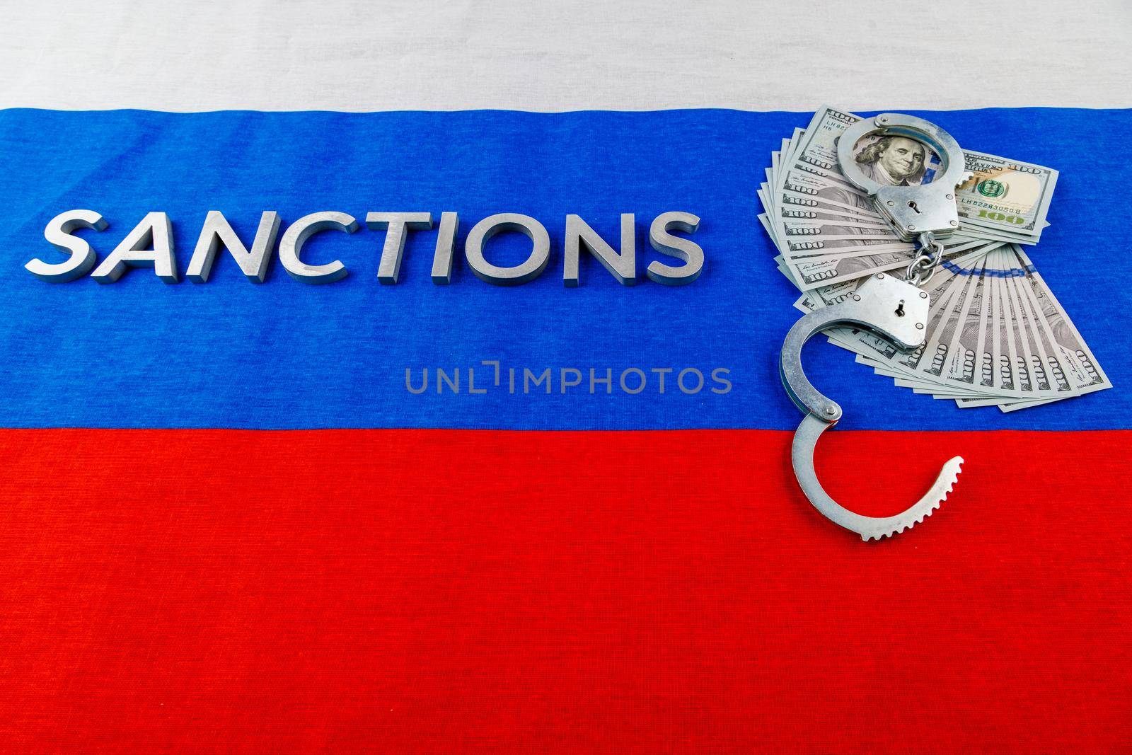 the word sanctions laid with silver metal letters on russian tricolor flag near dollar banknotes and handcuffs in linear perspective by z1b