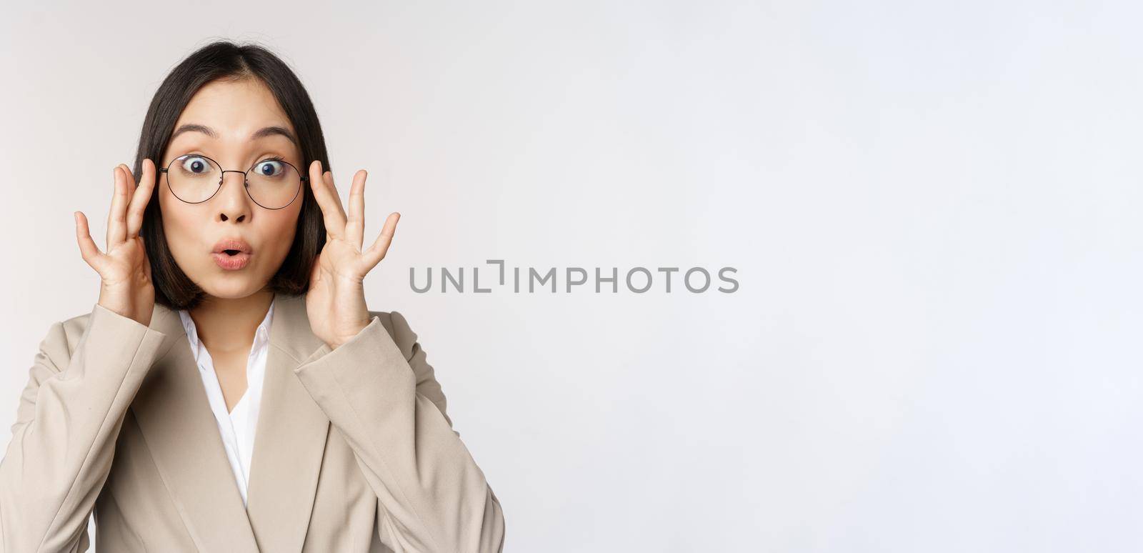 Portrait of asian businesswoman in glasses, looking surprised amazed at camera, standing in suit over white background by Benzoix