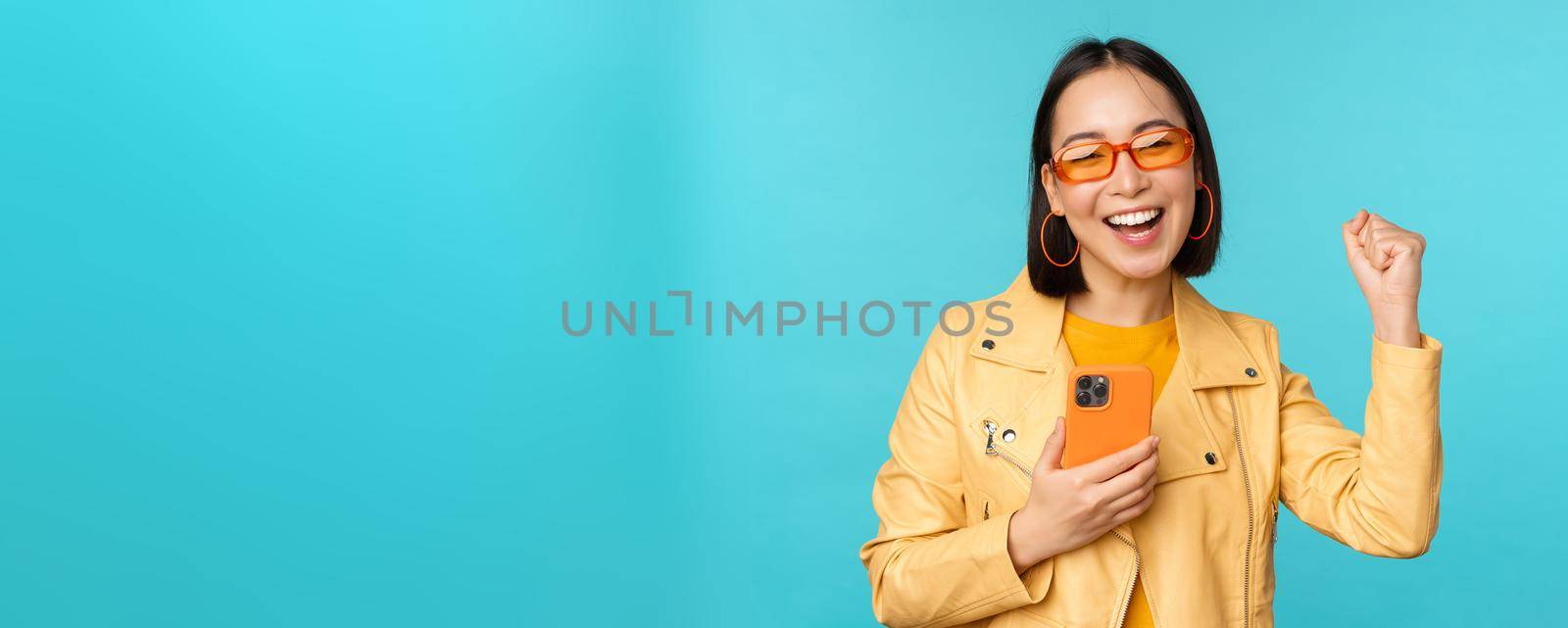 Enthusiastic smiling asian girl in sunglasses, holding mobile phone and dancing, laughing with smartphone, standing over blue background.