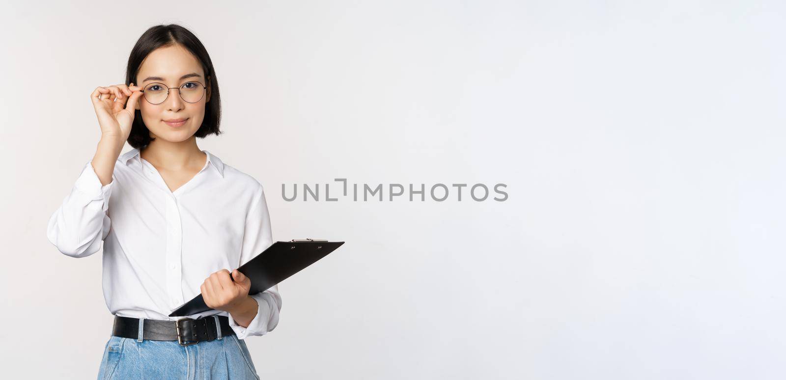 Young woman, office worker manager in glasses, holding clipboard and looking like professional, standing against white background.