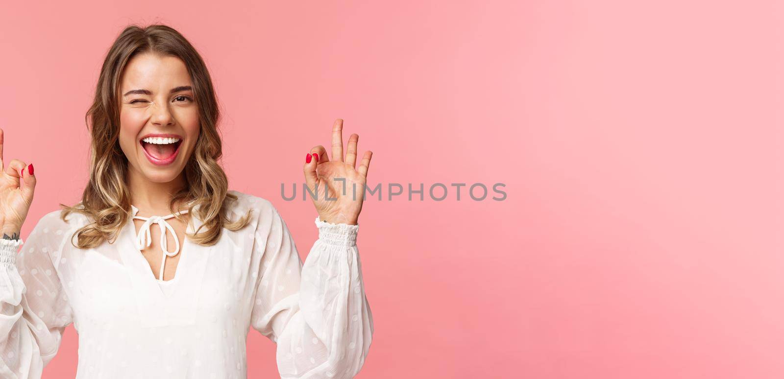 Close-up portrait of beautiful blond girl in white dress guarantee you will enjoy this event, show okay sign, confirm everything excellent, wink and smiling carefree, standing pink background.