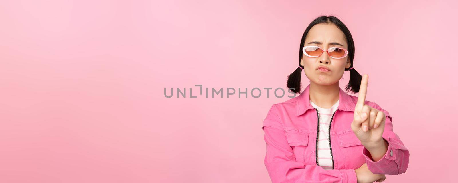 Image of serious, stylish asian girl in sunglasses, showing stop, prohibit gesture, taboo sign, saying no, standing over pink background.