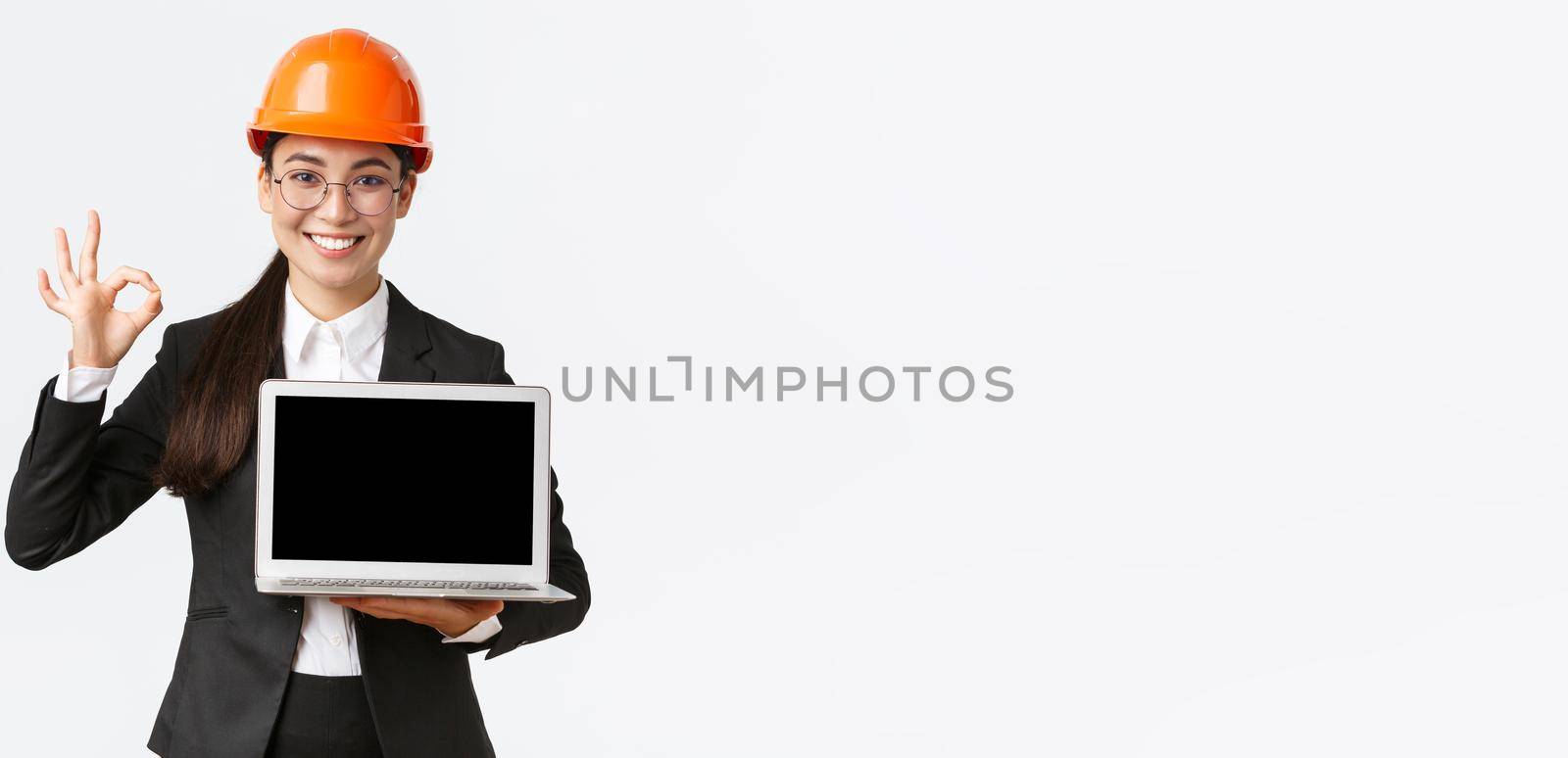 Smiling professional female architect, construction manager at factory showing graph, positive diagram, make okay gesture in approval and holding laptop facing screen, wear safety helmet by Benzoix