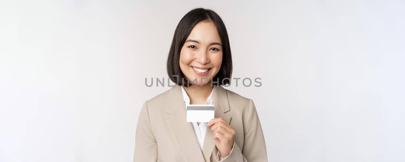 Smiling office clerk, asian corporate woman showing credit card, standing over white background in beige suit. Copy space