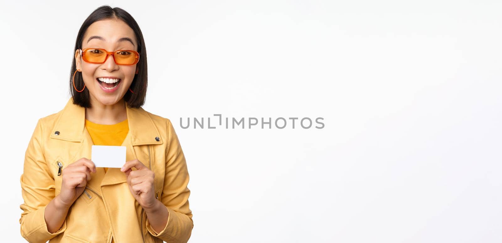 Portrait of beautiful modern asian girl in sunglasses, smiling happy, showing credit card, standing over white background. copy space