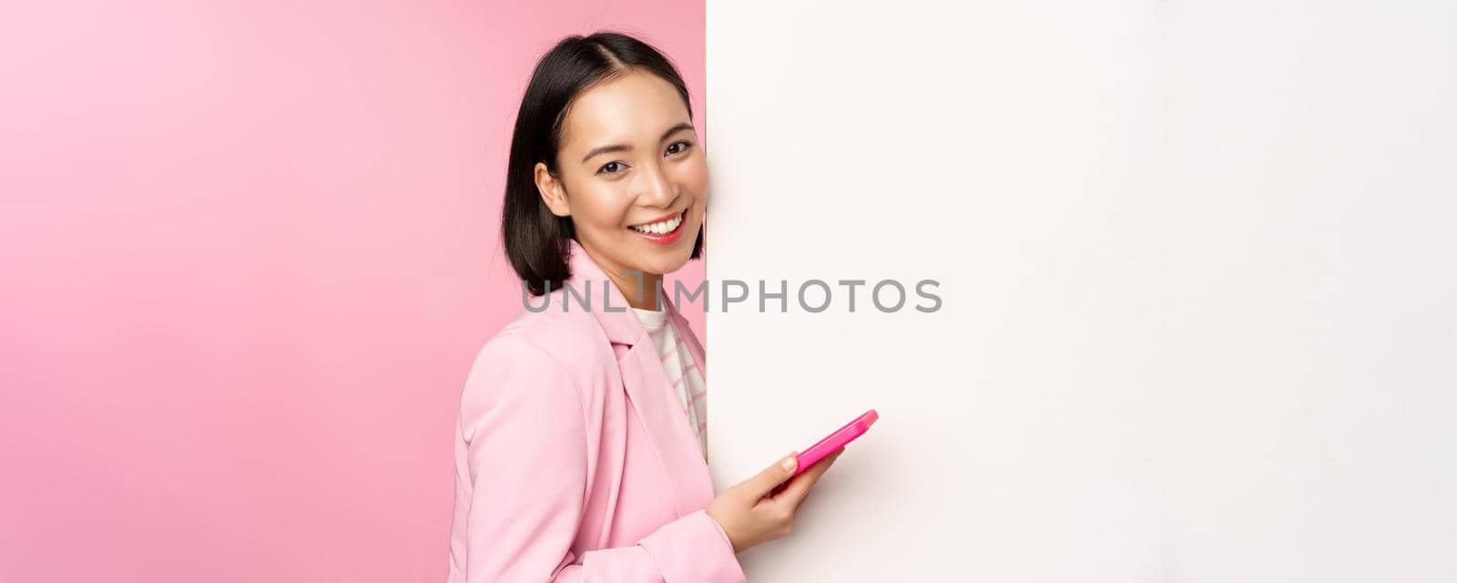 Image of korean female entrepreneur in suit, standing near info wall, advertisement on board, holding smartphone and smiling, posing over pink background by Benzoix