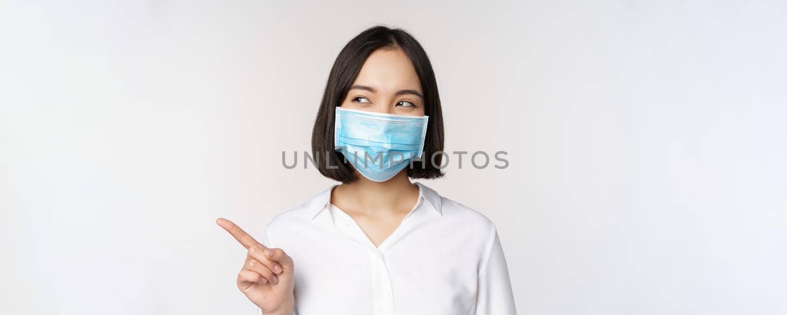 Portrait of cute asian woman in medical face mask, coronavirus protection, pointing finger left and looking intrigued at empty copy space, white background.