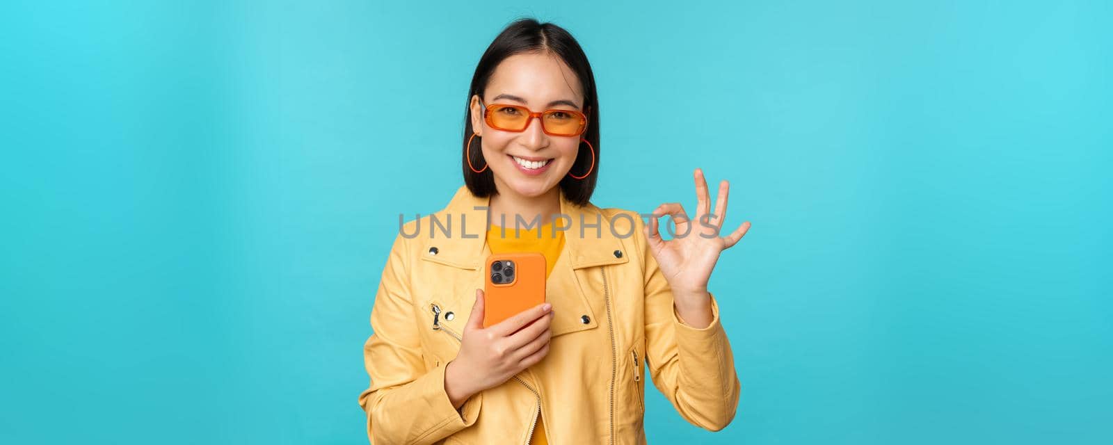 Smiling asian girl with smartphone, shwoing okay, ok sign in approval, standing over blue background. Copy space