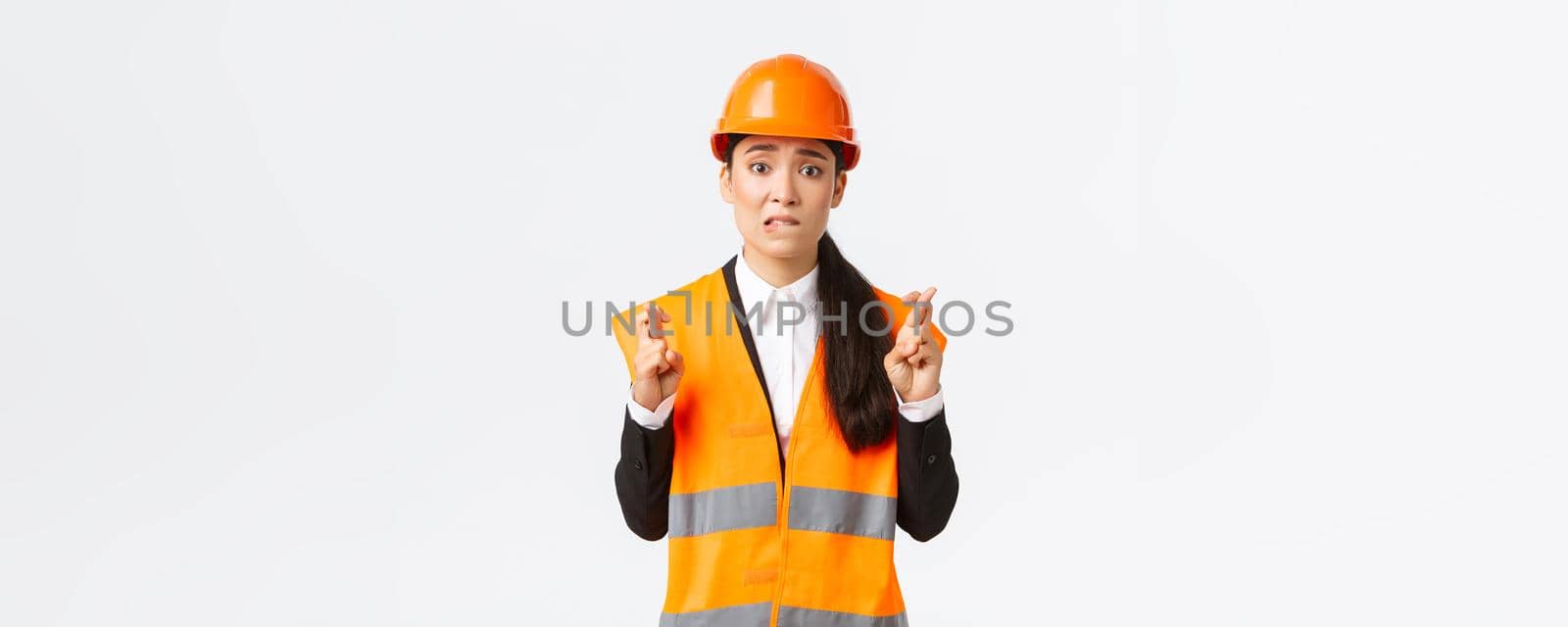 Worried hopeful asian female architect feeling nervous as waiting for inspection results at construction area, wearing safety helmet, cross fingers good luck, hope everything be well by Benzoix