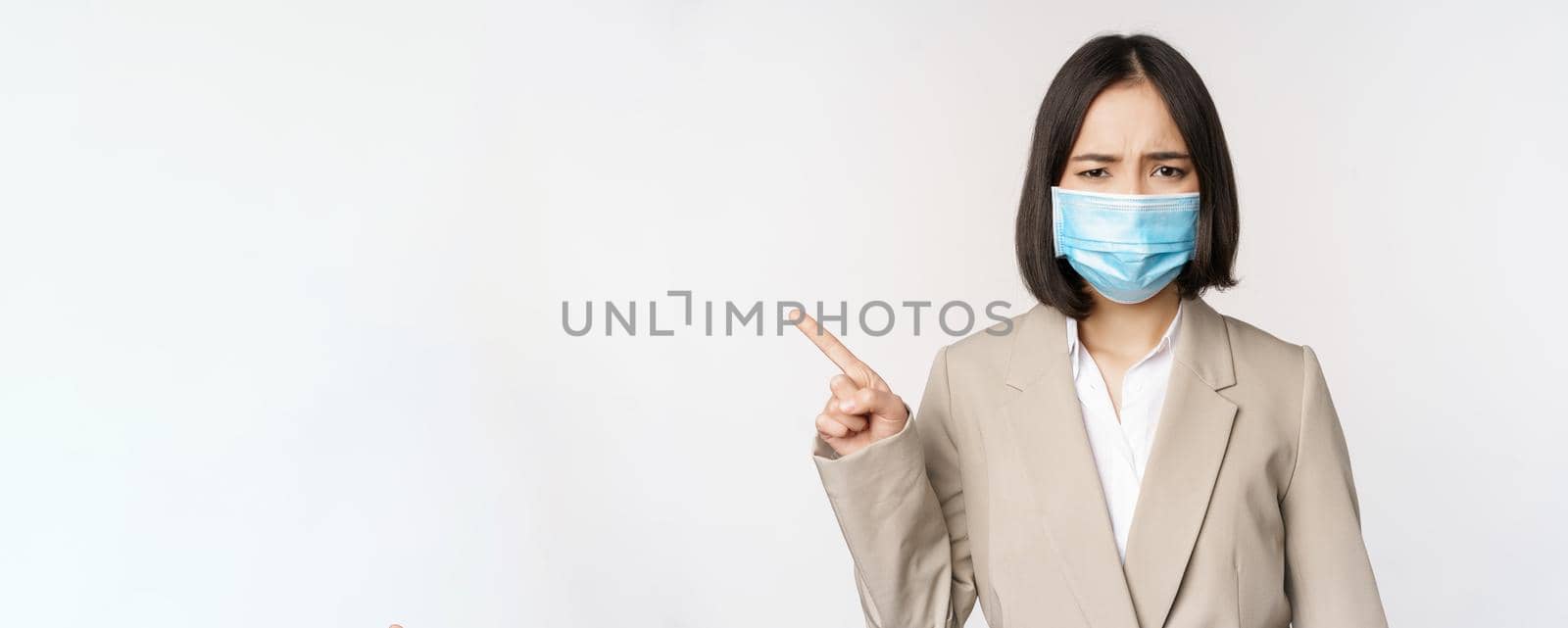 Coronavirus and work concept. Frustrated asian business woman in medical face mask, pointing finger left and complaining, standing over white background by Benzoix