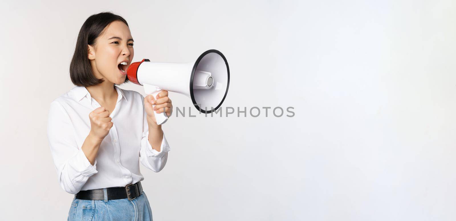 Enthusiastic asian woman, girl activist shouting at protest, using megaphone, looking confident, talking in loudspeaker, protesting, standing over white background by Benzoix