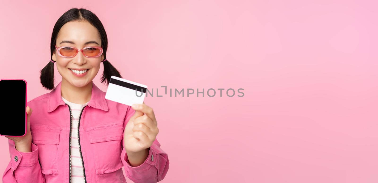 Image of smiling korean woman showing credit card and mobile phone screen, smartphone application interface, paying online, shopping contactless, standing over pink background.