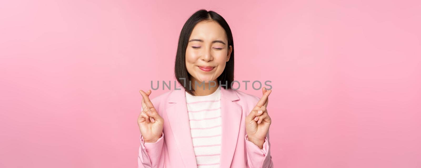Happy lucky businesswoman, asian corporate lady wishing, making wish, hoping for smth and praying, standing in suit over pink background.