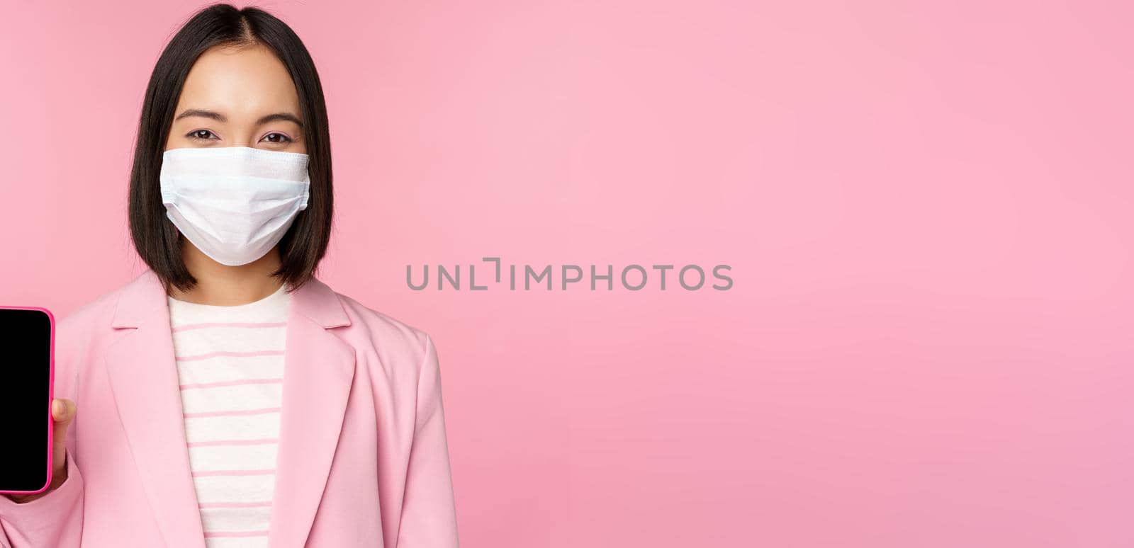 Portrait of smiling korean saleswoman in medical face mask, business suit, showing smartphone screen, standing over pink background.