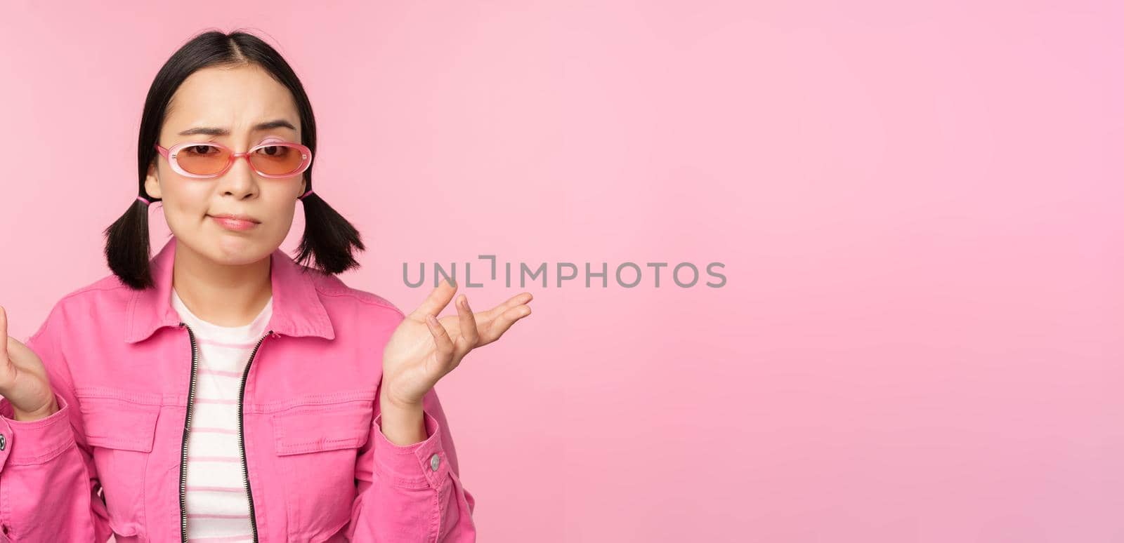 Close up portrait of asian girl looking confused, shrugging puzzled and looking at camera, wearing sunglasses, standing over pink background.