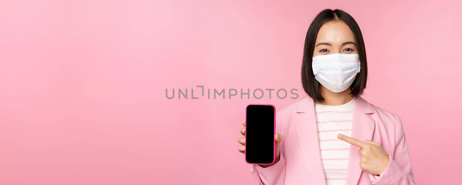 Smiling korean businesswoman in suit, wearing medical face mask from covid, pointing finger at mobile phone screen, standing over pink background.