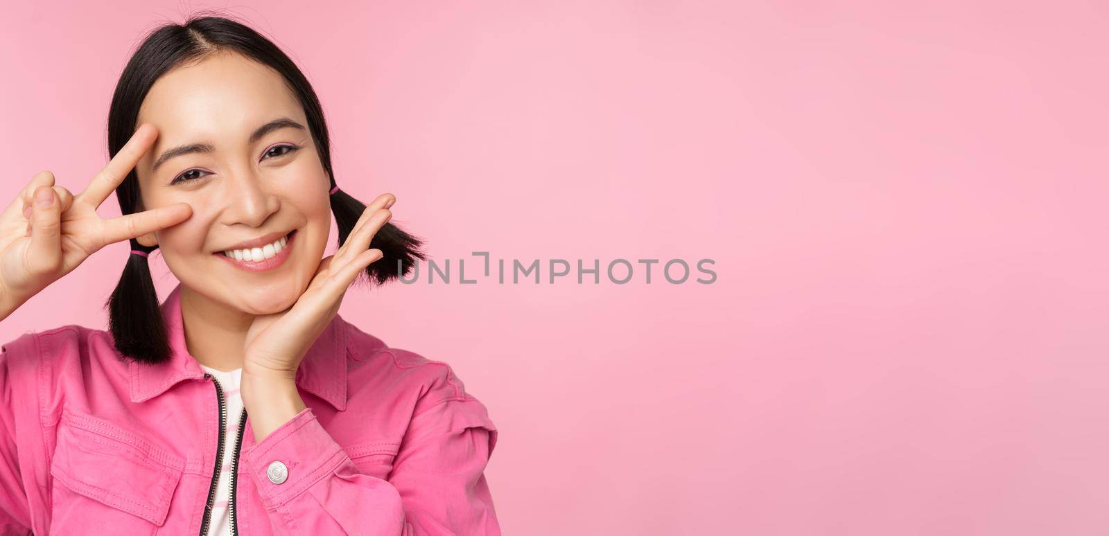 Close up of stylish asian girl smiles happy, shows peace v-sign, kawaii pose, posing against pink background. Copy space